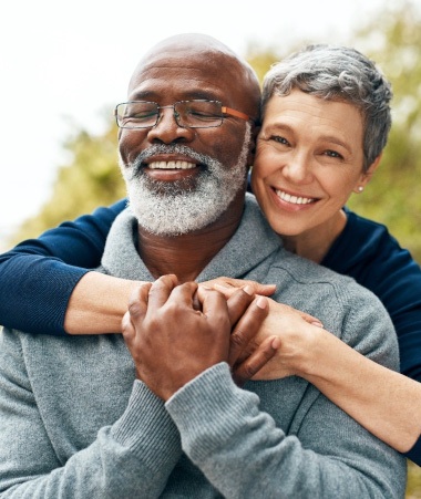 Woman in blue shirt outside hugging man in grey sweater from behind