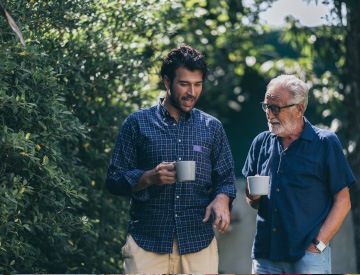 Younger man in blue checkered shirt talking with older man in blue t-shirt outside over coffee