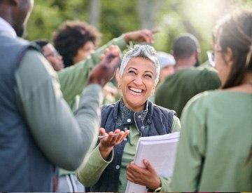 Woman with short grey hair in green shirt outside with notebook and pen working a crowd of people